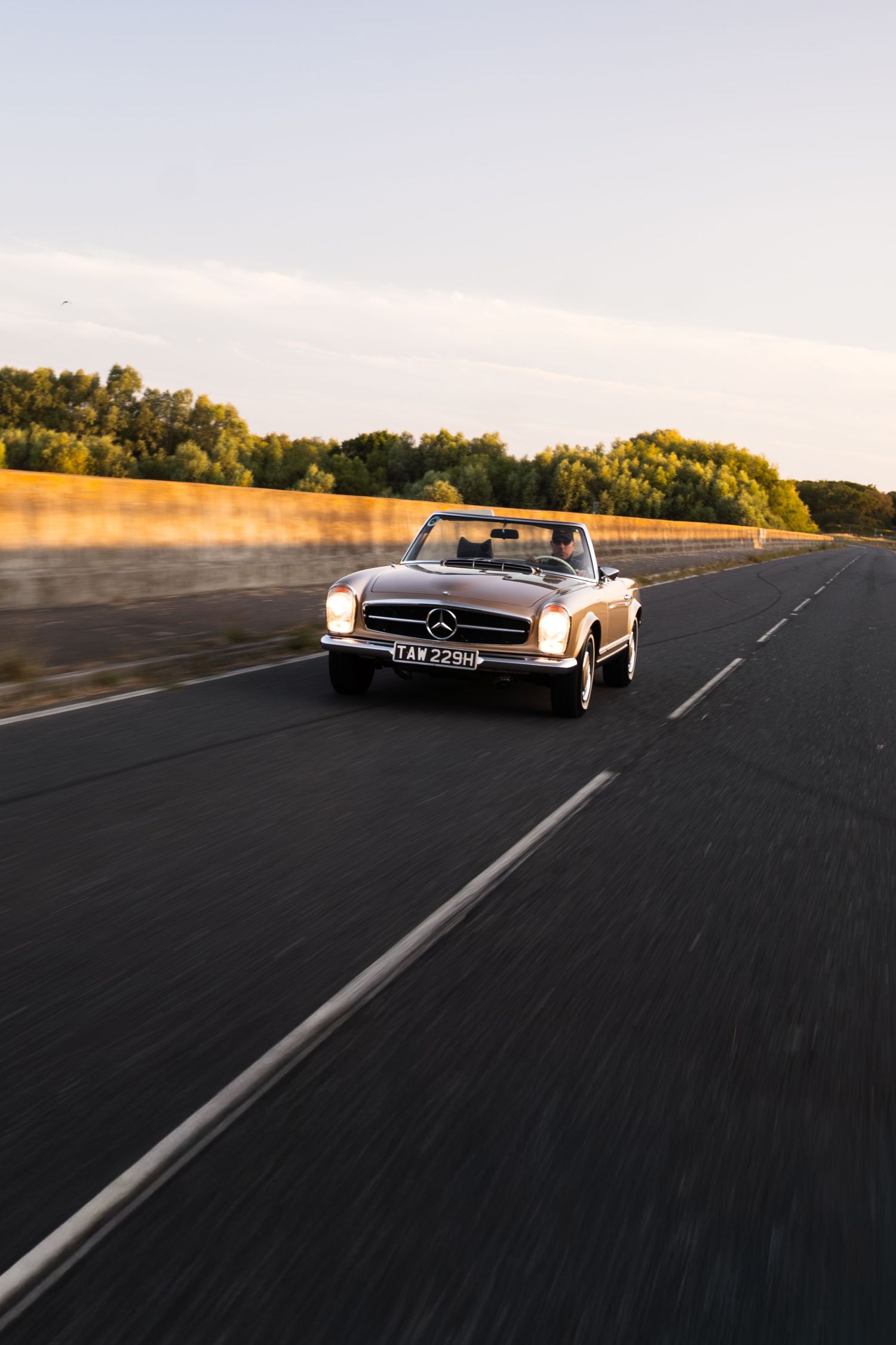 Classic Mercedes-Benz convertible driving on an open road at golden hour with trees in the background.