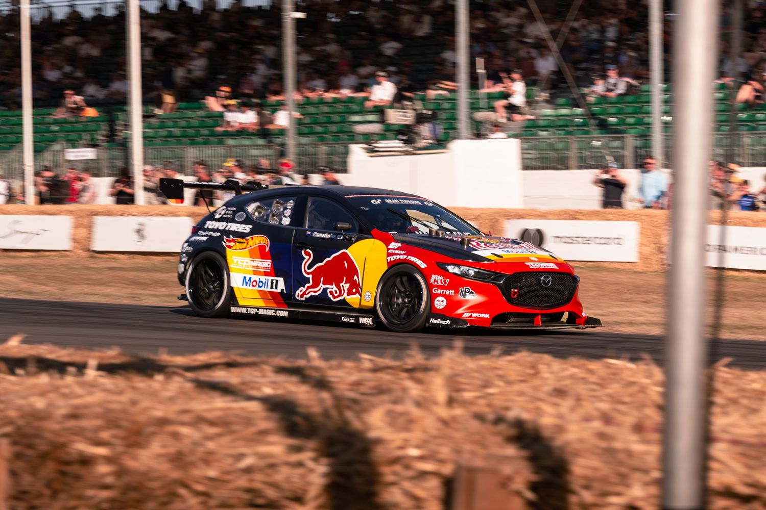 Red Bull rally car racing on a dirt track at a motorsport event, with spectators watching in the background.