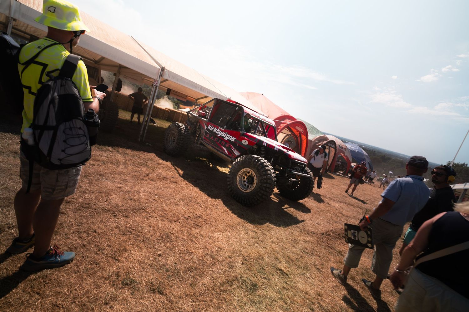 Off-road race vehicle at outdoor rally event with spectators and green tent in dusty field on sunny day.