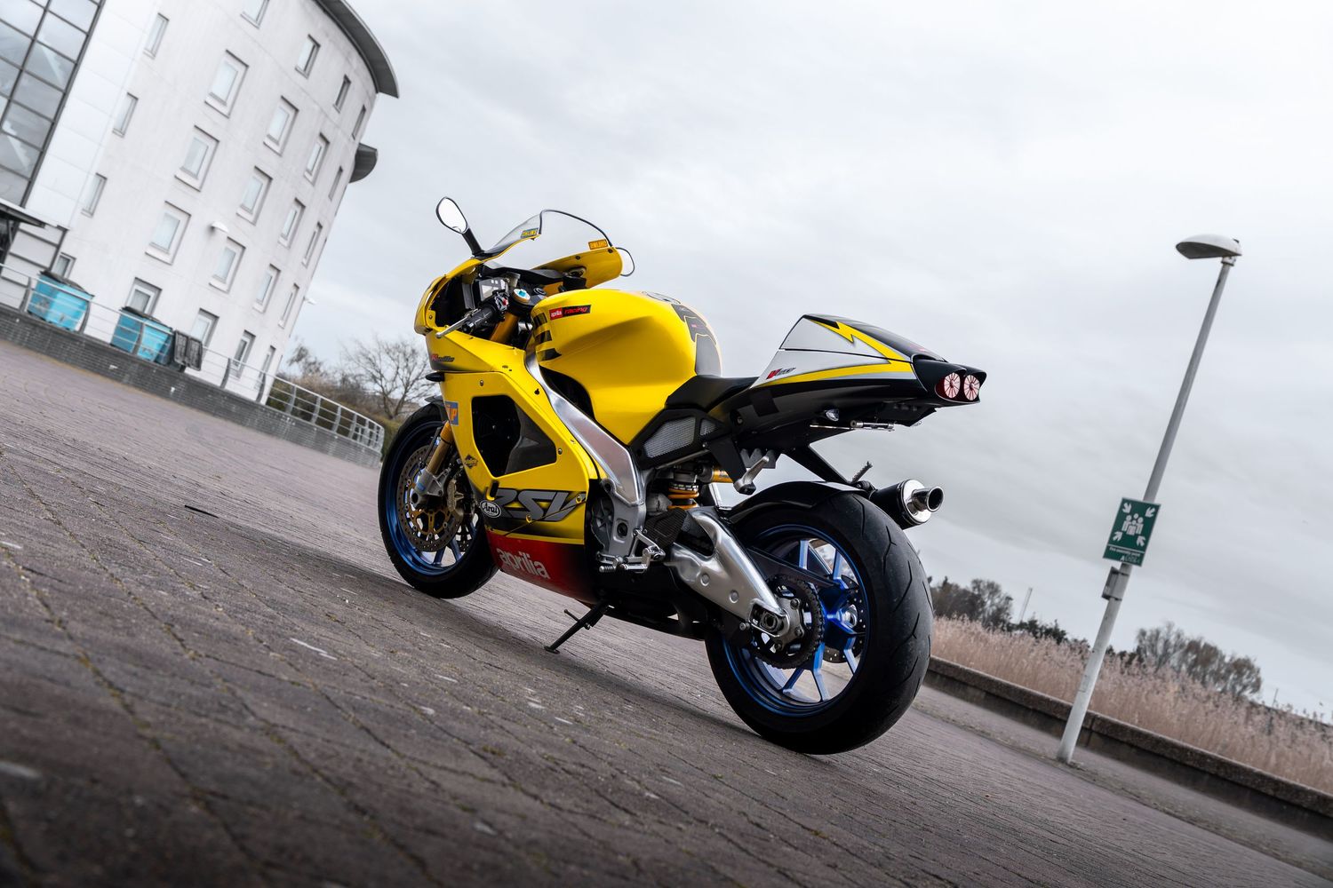Yellow Aprilia RSV motorcycle shot from low rear angle against a cloudy sky and modern building.