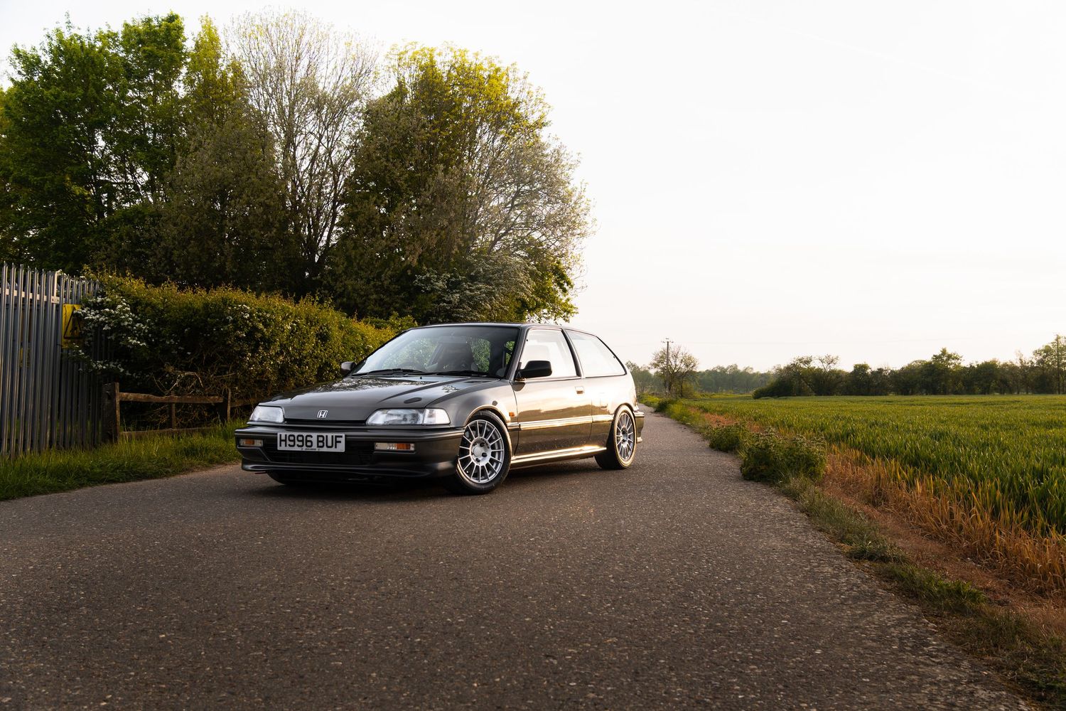 Three-quarter front view of a lowered grey Honda Civic on a rural road surrounded by trees and fields.