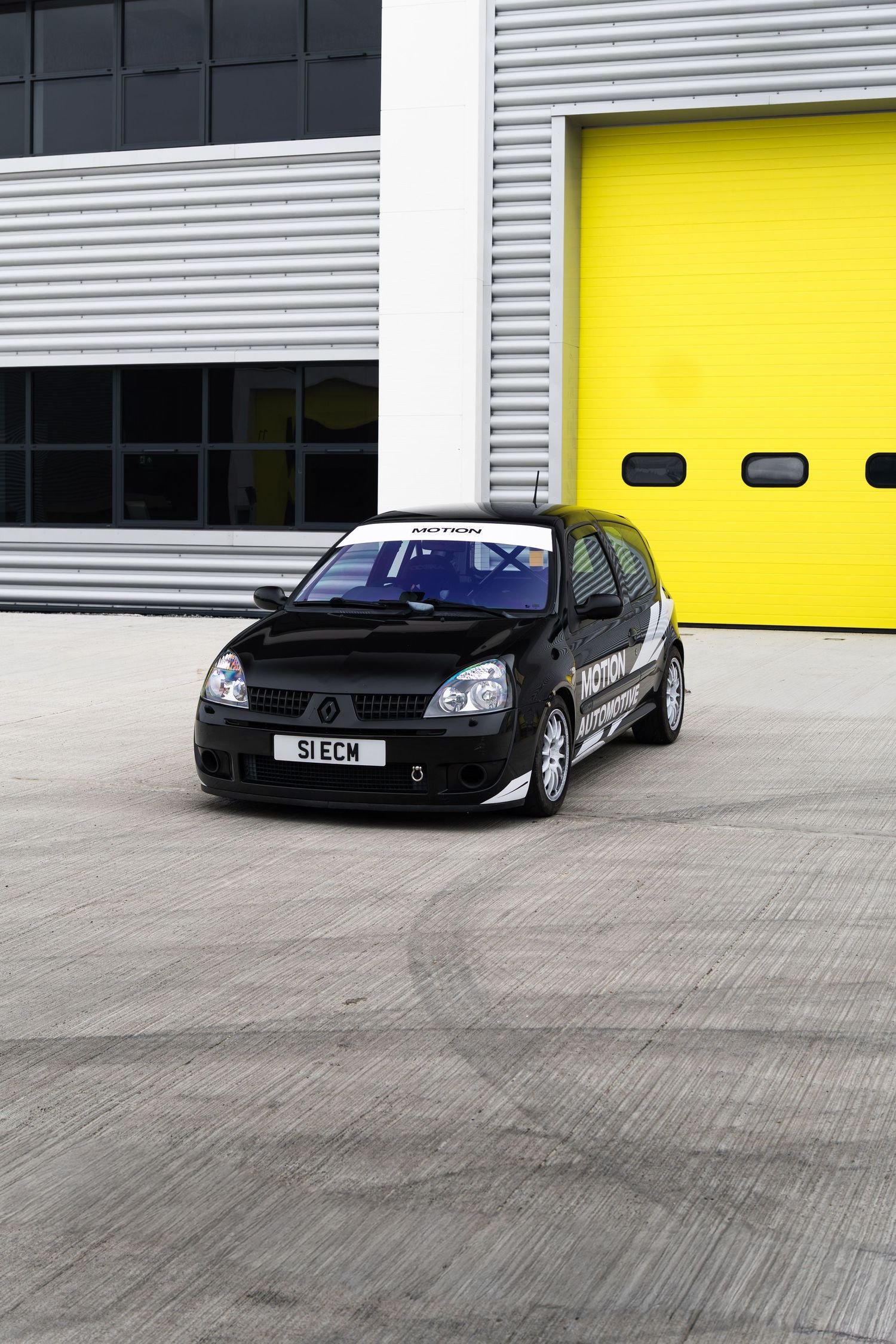 Black Renault Clio race car with Motion livery parked outside industrial unit with yellow doors.