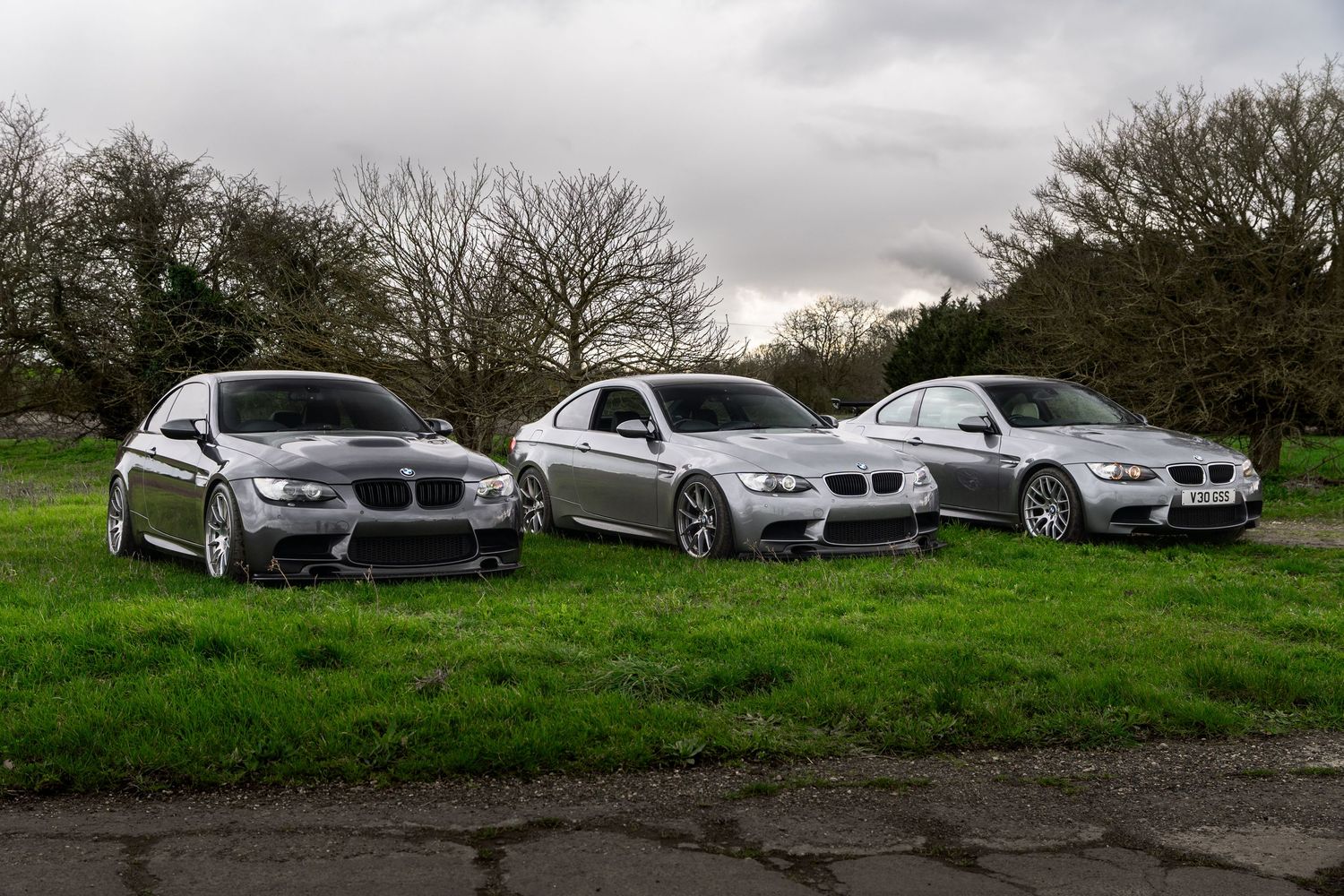 Three silver BMW M3 coupes parked on grass in a countryside setting under a cloudy sky.