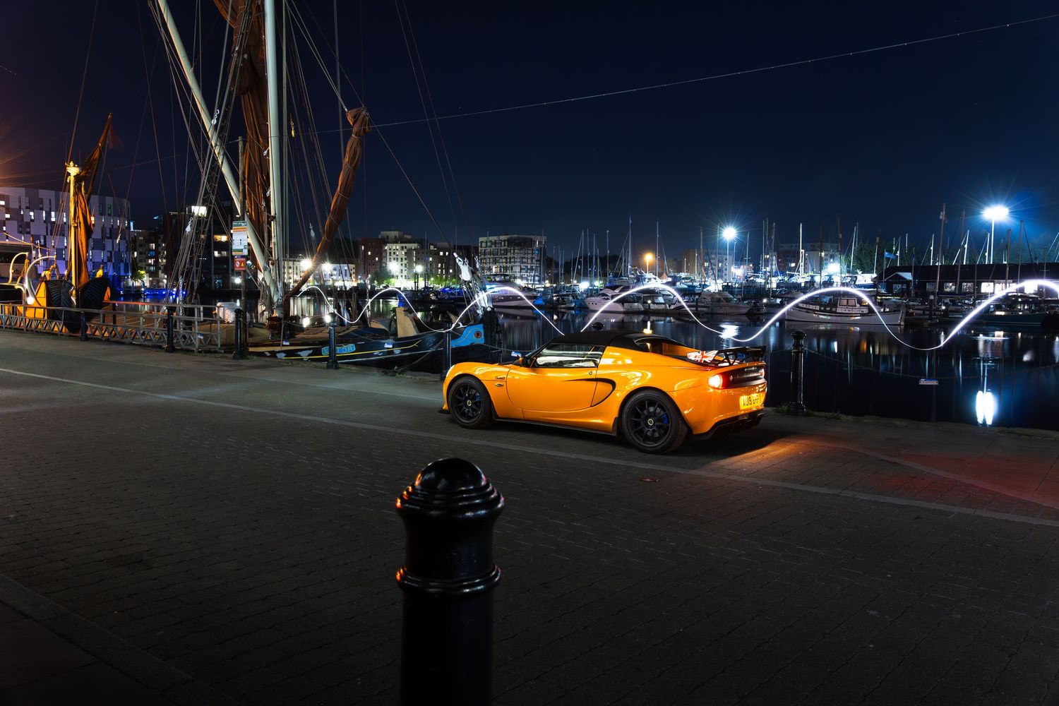 Yellow Lotus Elise sports car at a busy marina at night with a fire hydrant in the foreground.