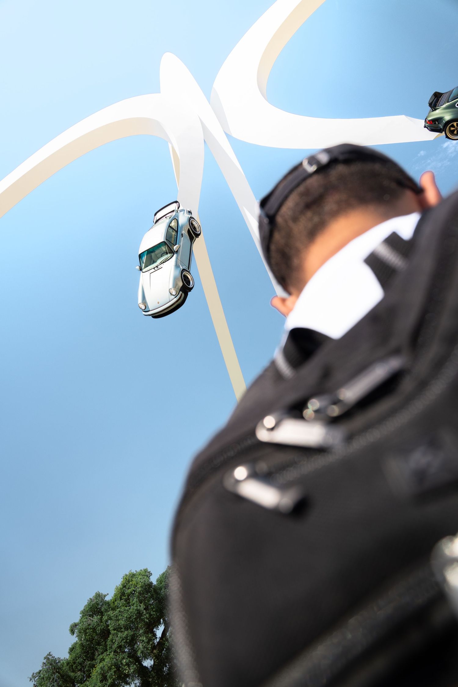Low-angle view of a man in black jacket with toy cars on a white track against a blue sky.