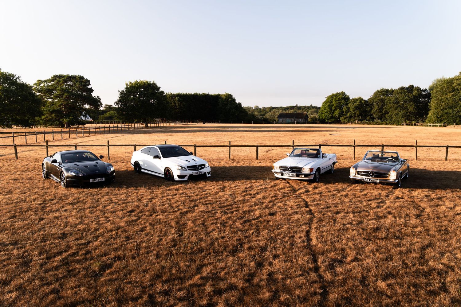 Four luxury Mercedes-Benz cars parked in a row on a golden field with wooden fencing and trees in background.