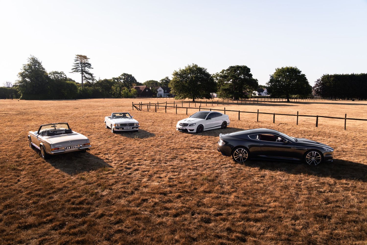 Four classic and modern Aston Martin cars parked in a golden field with trees and fence in background.