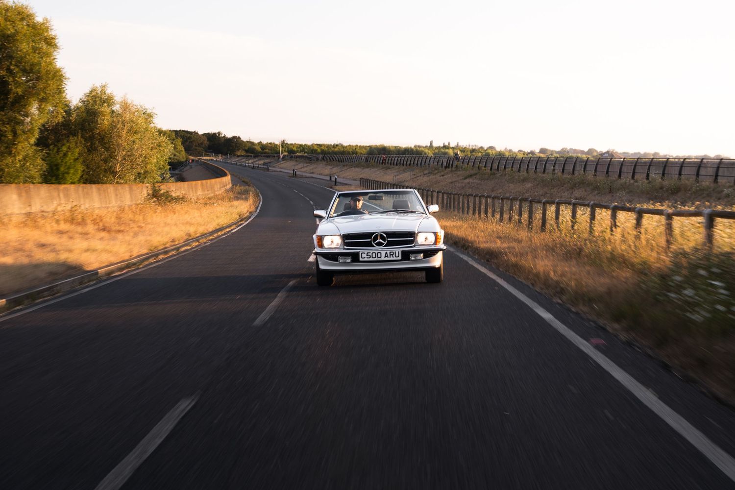 White classic Mercedes-Benz convertible driving on a rural road at golden hour, surrounded by trees and open landscape.