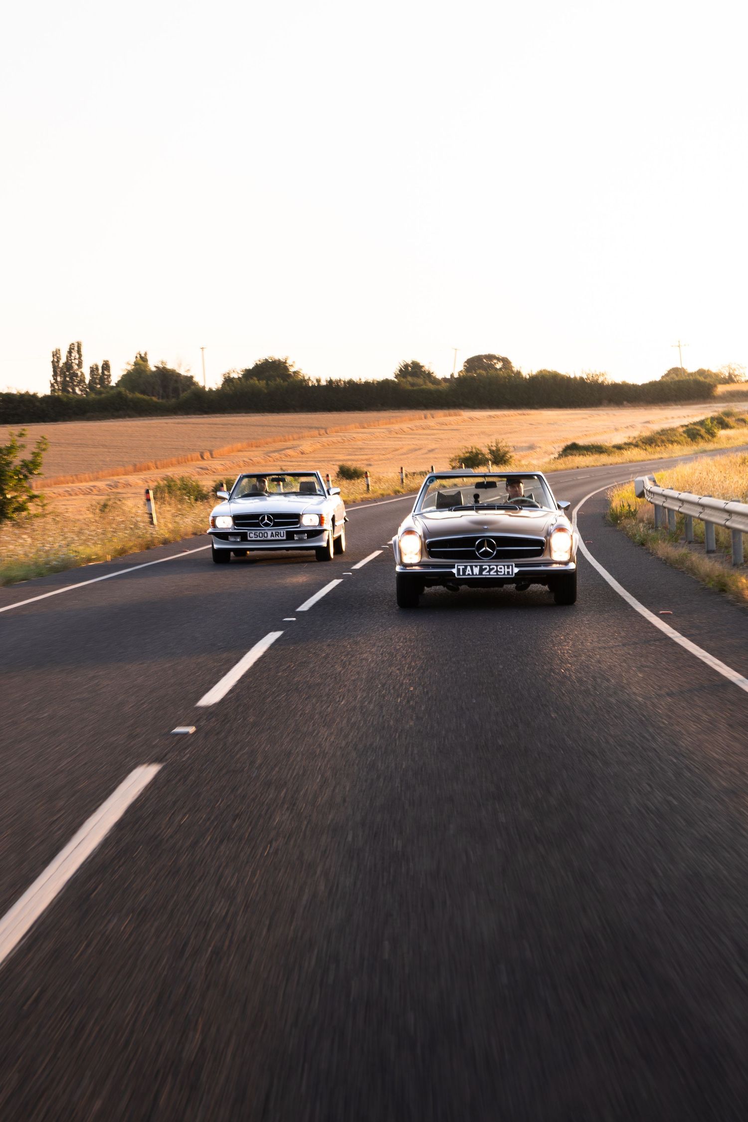 Two classic white Mercedes-Benz convertibles driving side by side on a rural road at golden hour sunset.