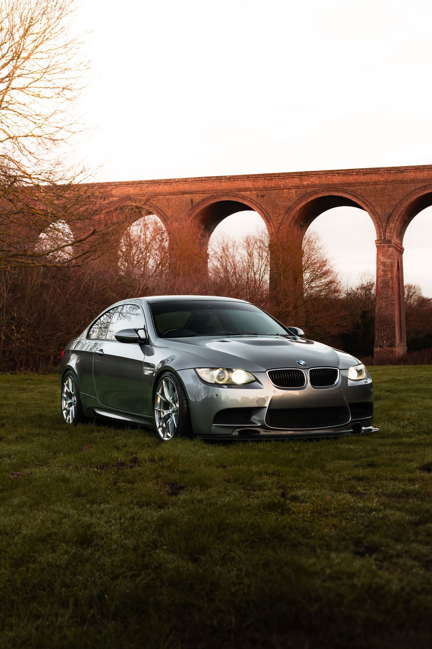 A silver BMW M3 parked on grass in front of a historic brick viaduct at dusk.