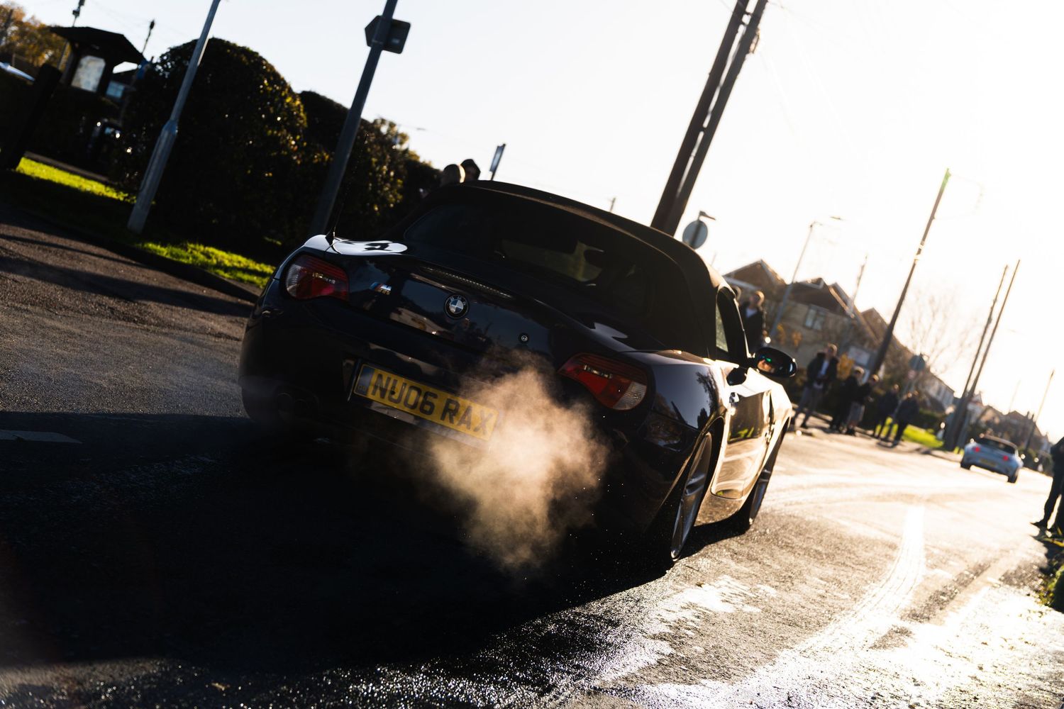 Black BMW car emitting exhaust smoke on a sunny street during a car meet event.