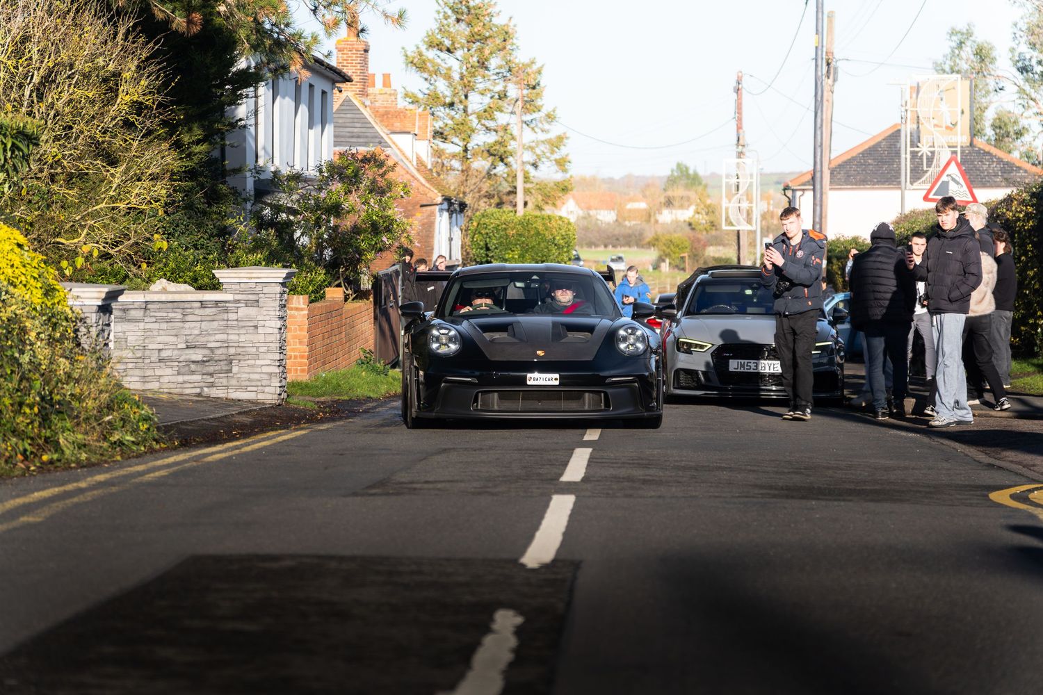 Black sports cars driving down a residential road surrounded by car enthusiasts at a meet.
