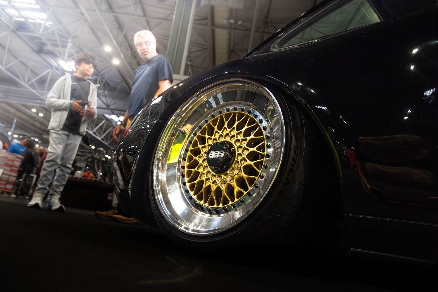 Close-up of a gold BBS mesh wheel on a black car at an auto show with spectators in background.