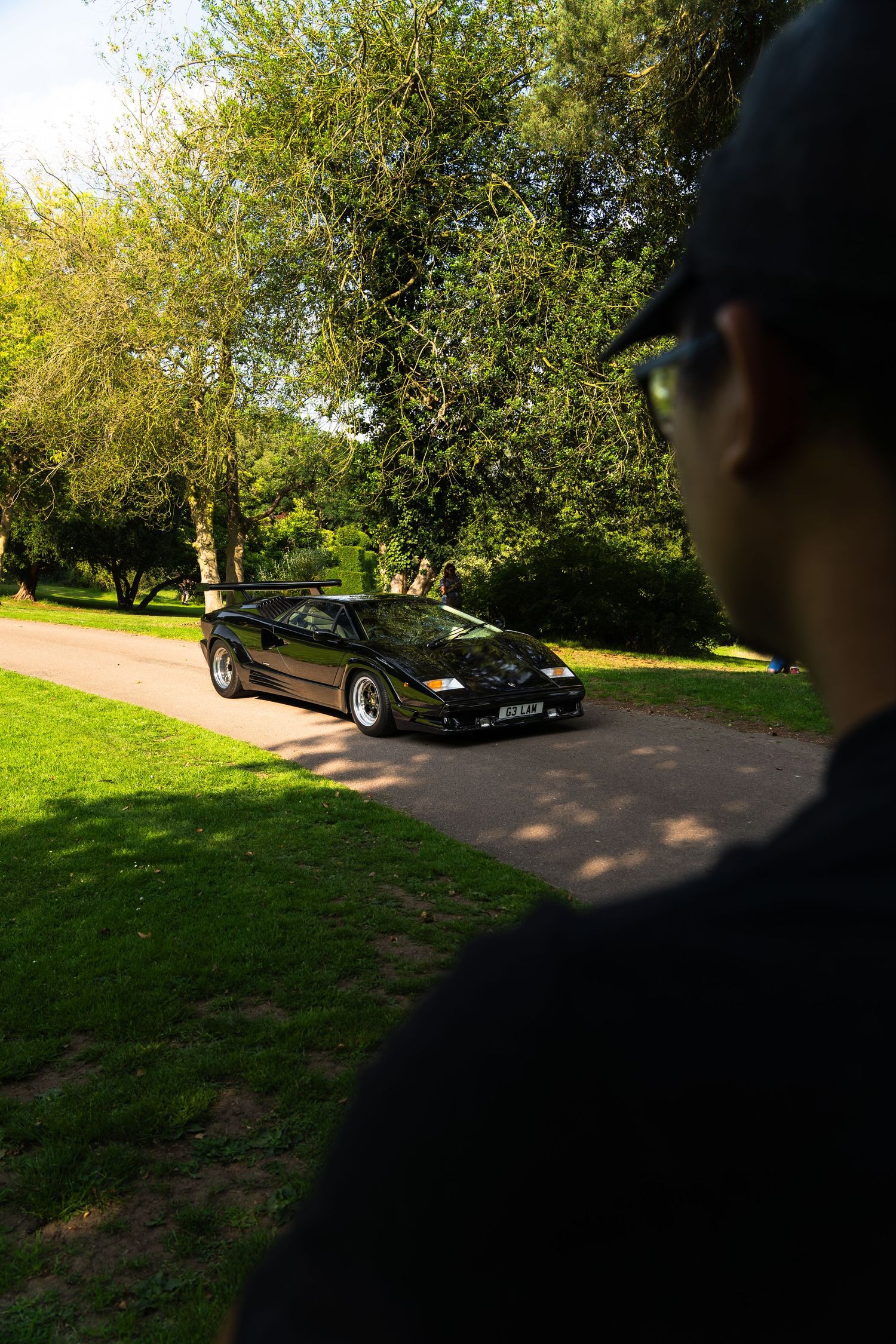 A man in a black cap admires a sleek black Lamborghini Countach parked on a sunlit path in a lush green park.