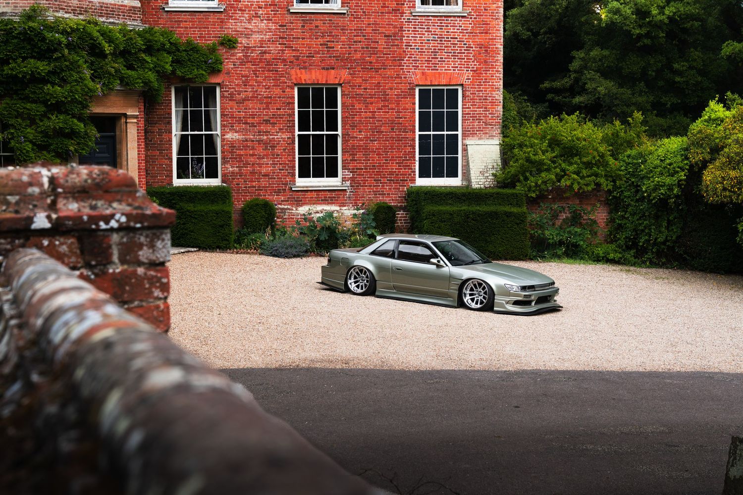 A lowered silver Nissan Silvia S14 parked on gravel outside a historic red brick English manor house.