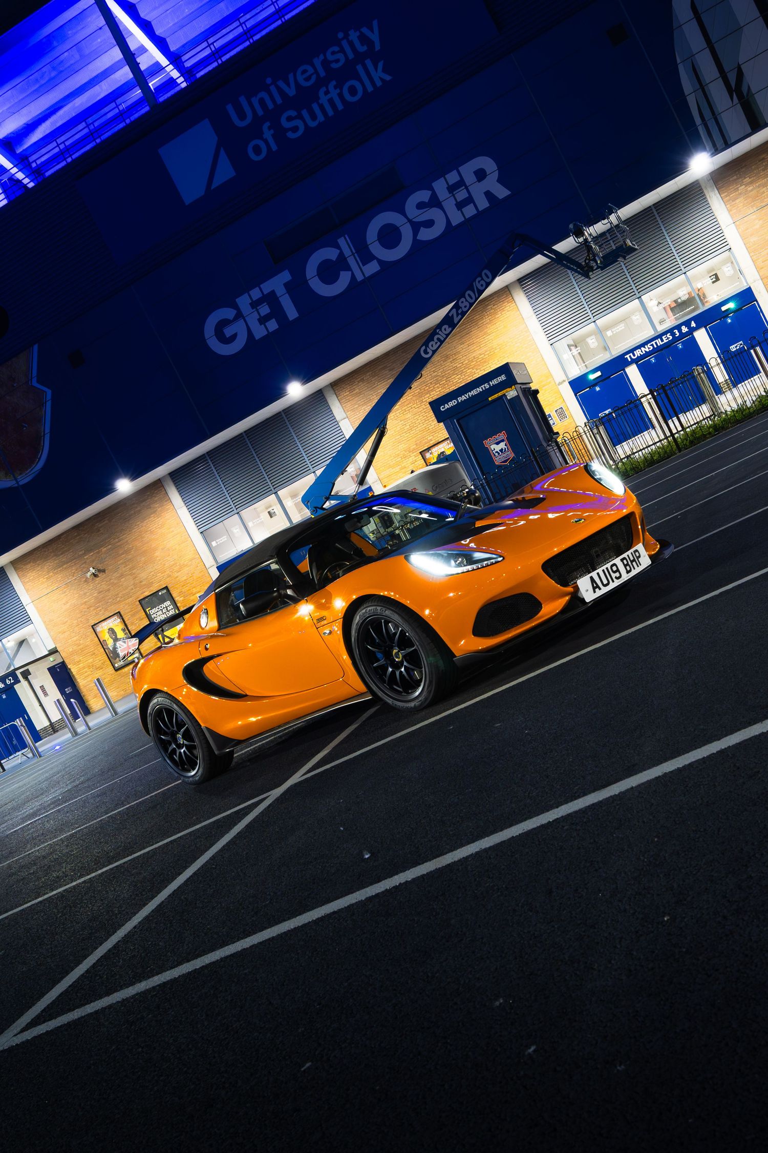 Orange Lotus Elise with open door parked outside University of Suffolk GET CLOSER stadium entrance at night.
