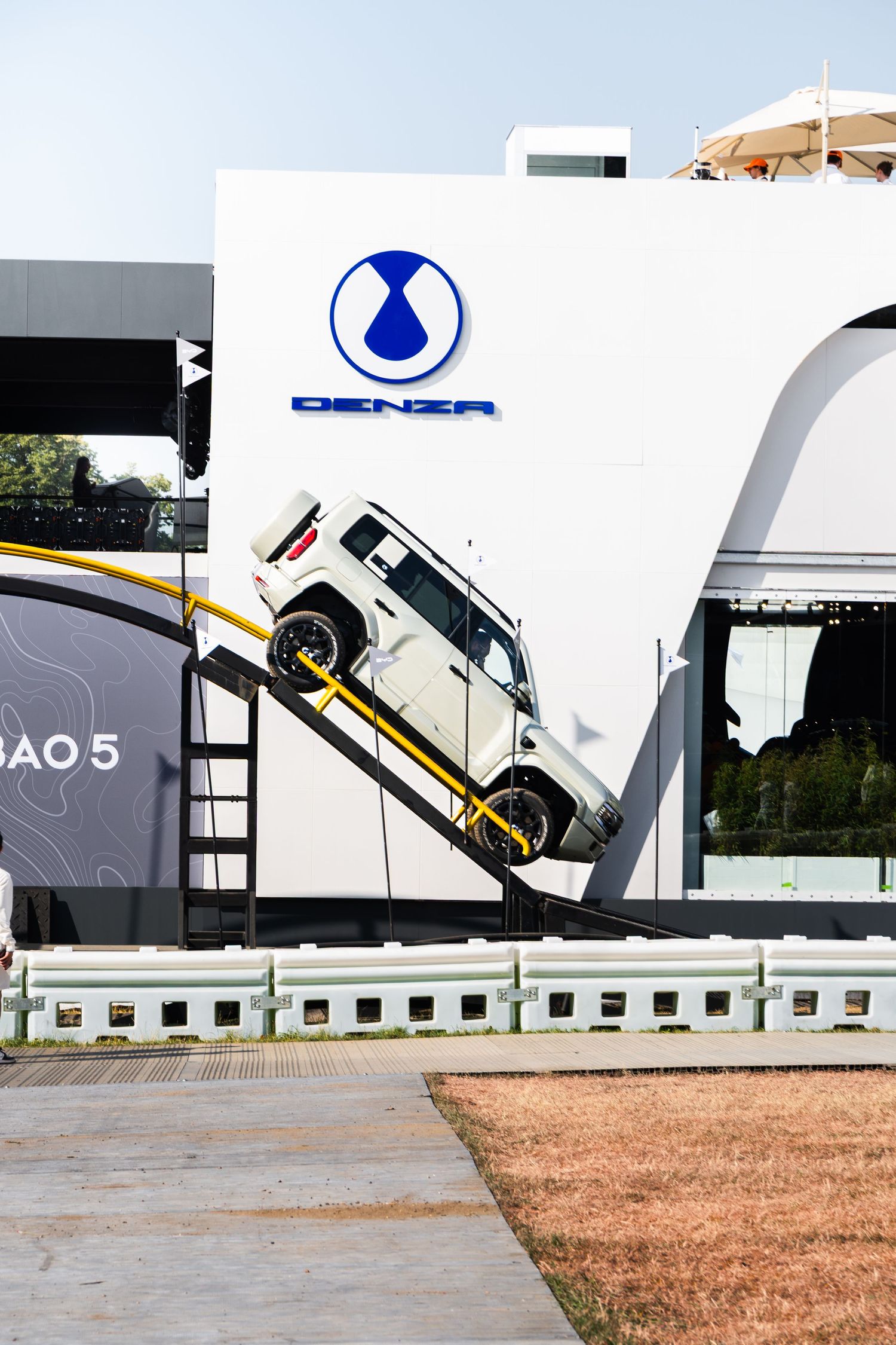 DENZA electric vehicle display at an outdoor auto show featuring a white SUV on an angled ramp beside a branded white wall.