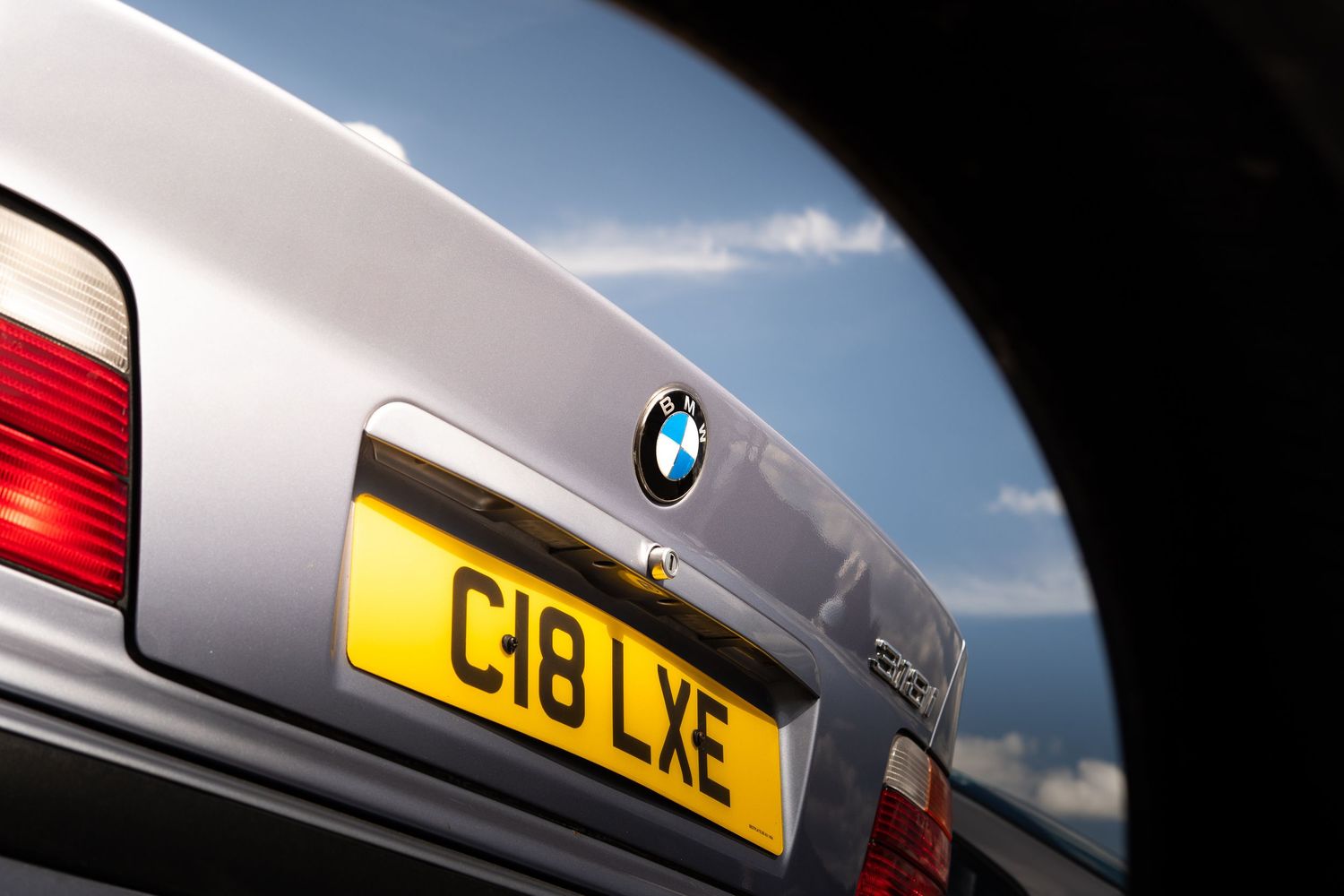 Rear view of a silver BMW with yellow UK license plate C18 LXE against a blue sky.