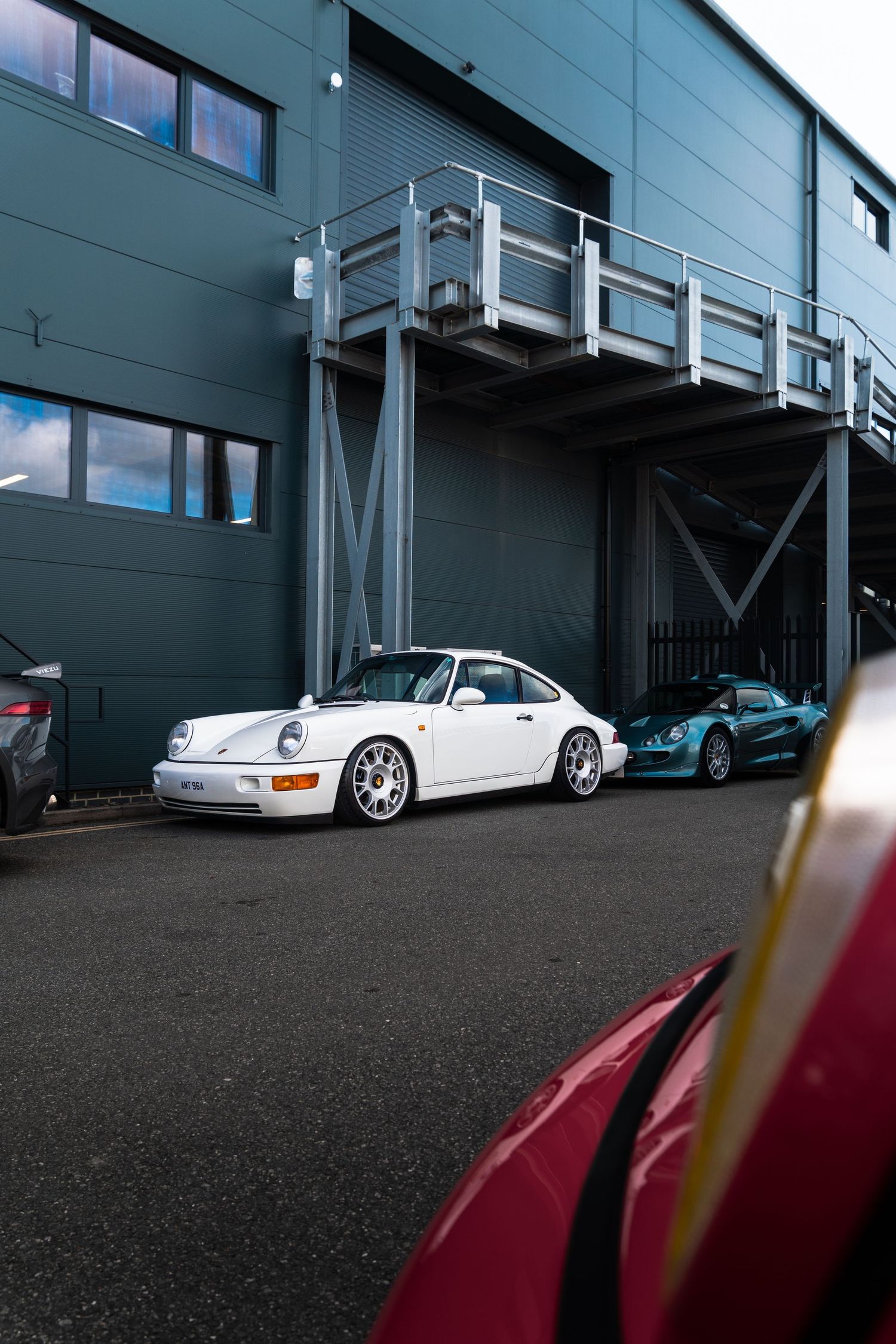 White Porsche 911 and teal sports car parked outside a grey industrial building with metal walkway.