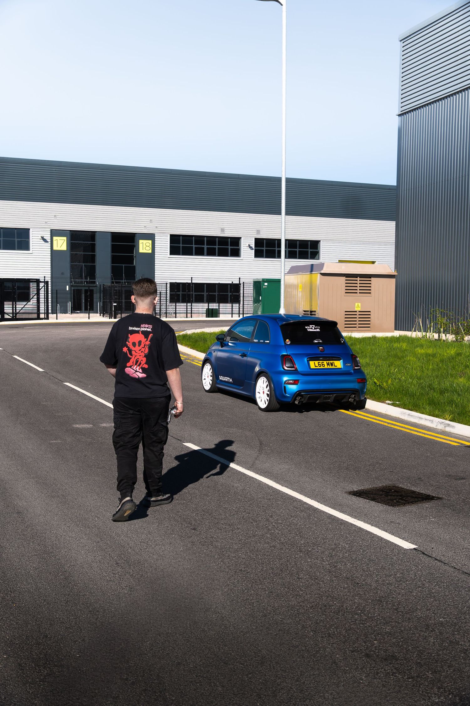Man walking away from a blue Abarth 500 on a road outside a modern industrial unit on a clear day.