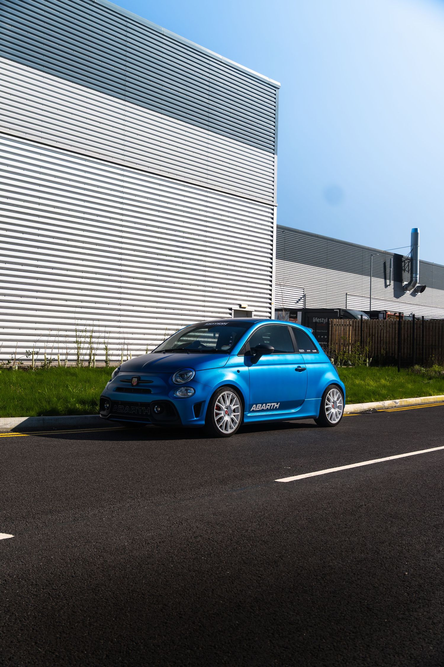 Blue Abarth 500 modified hatchback parked on a road beside a modern industrial building on a sunny day.