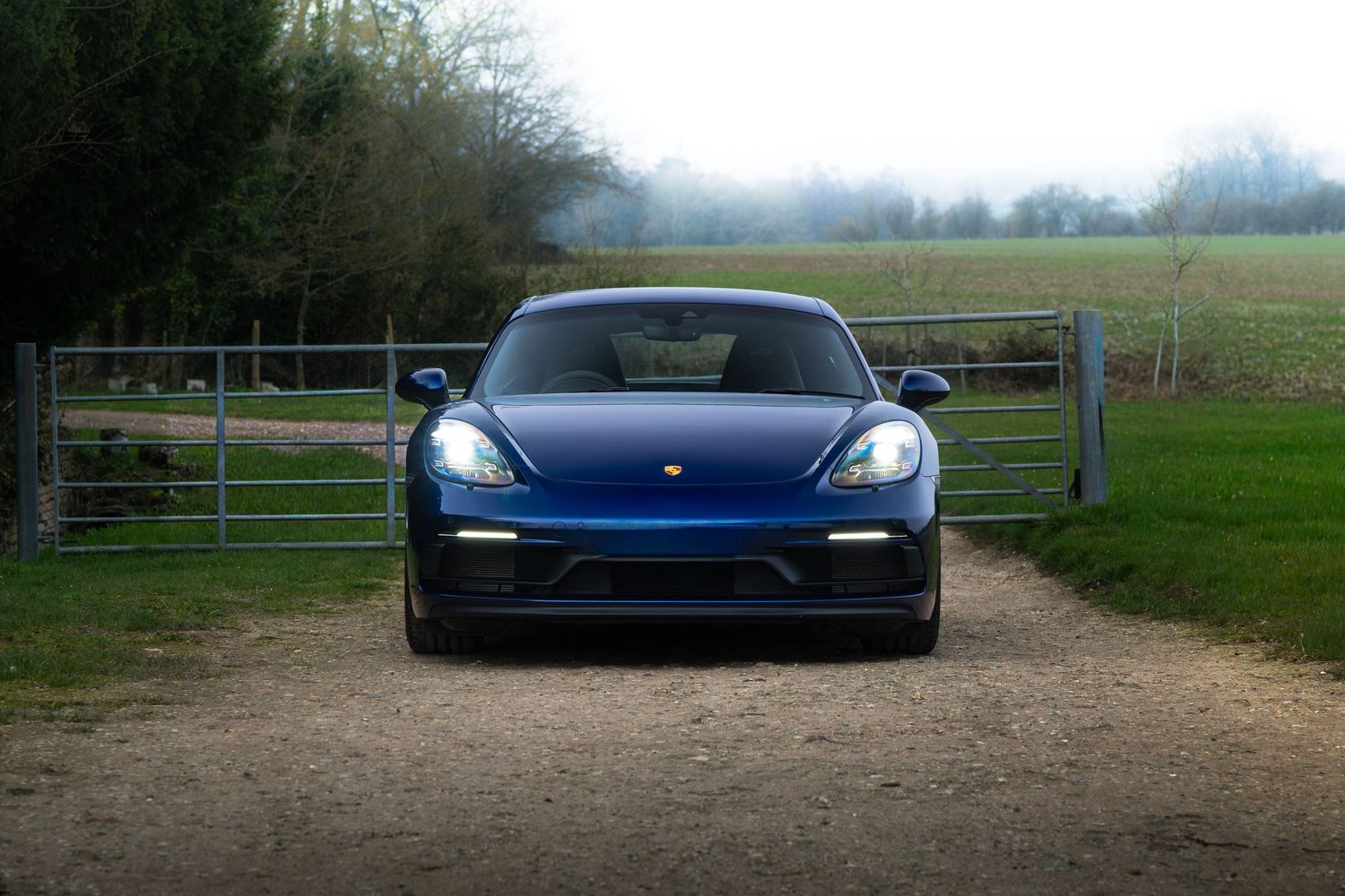 Front view of blue Porsche 718 Cayman GTS with headlights on, parked on rural gravel driveway near gate.