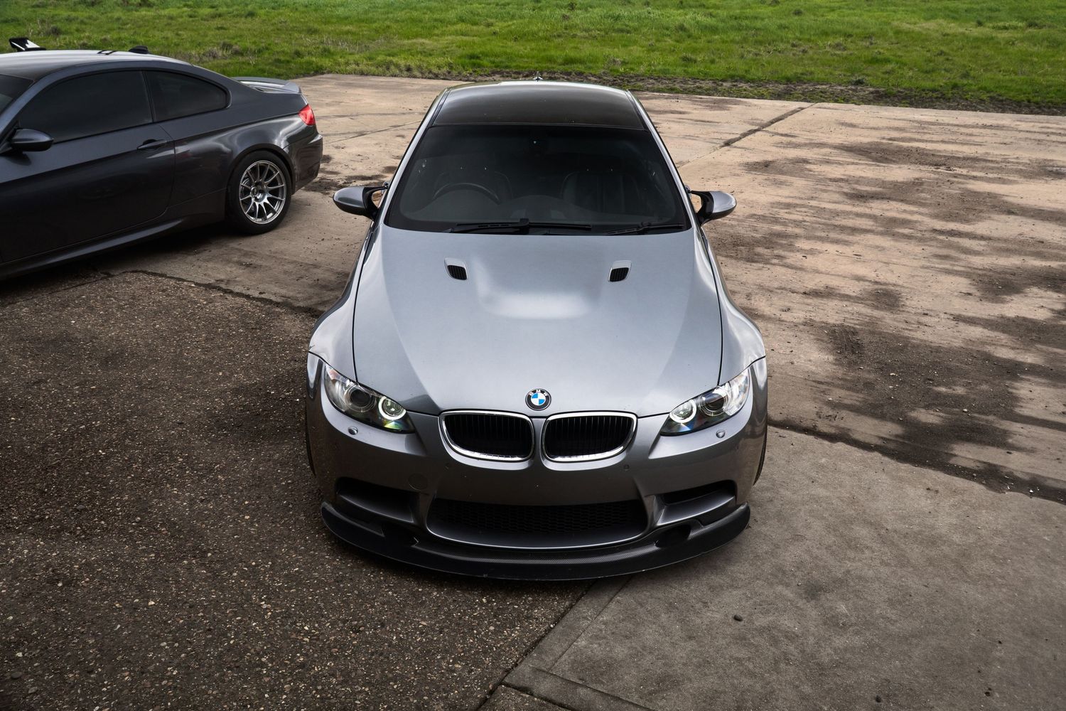 Front view of a silver BMW M3 with carbon fiber splitter and hood vents on wet pavement.