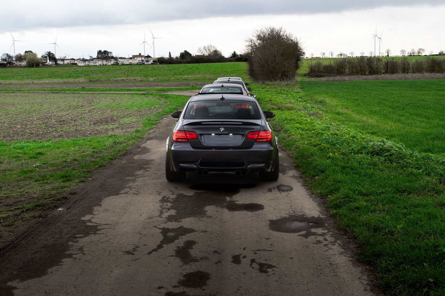 Black BMW M3 coupe viewed from rear on rural dirt path surrounded by green fields.