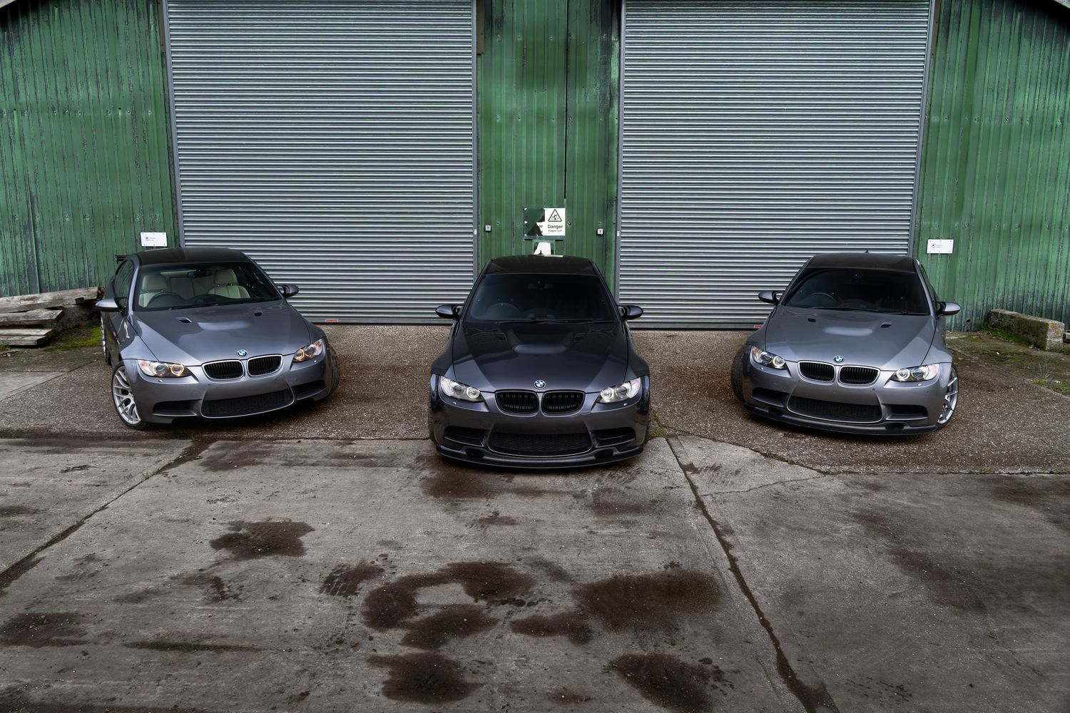Three grey BMW M3 models lined up outside a green corrugated metal warehouse.