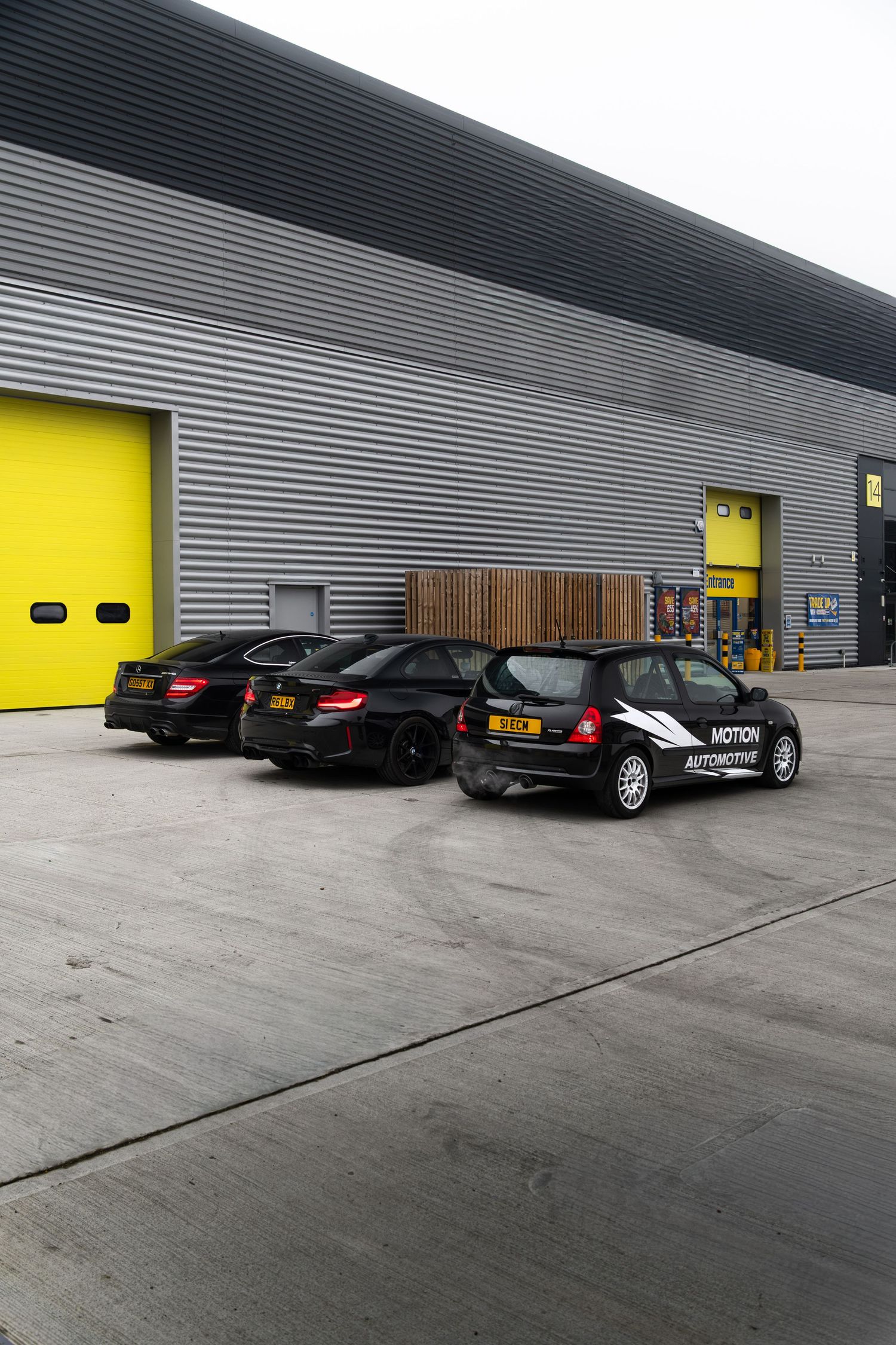 Rear view of three black Motion Automotive cars parked outside an industrial unit with yellow doors.