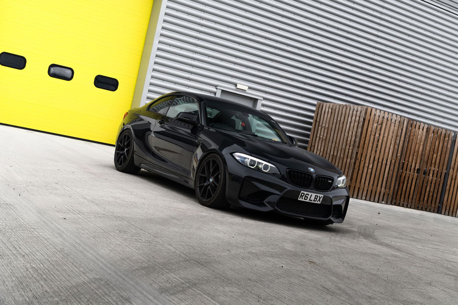 Black BMW M2 coupe parked outside a building with yellow garage doors and wooden fence.