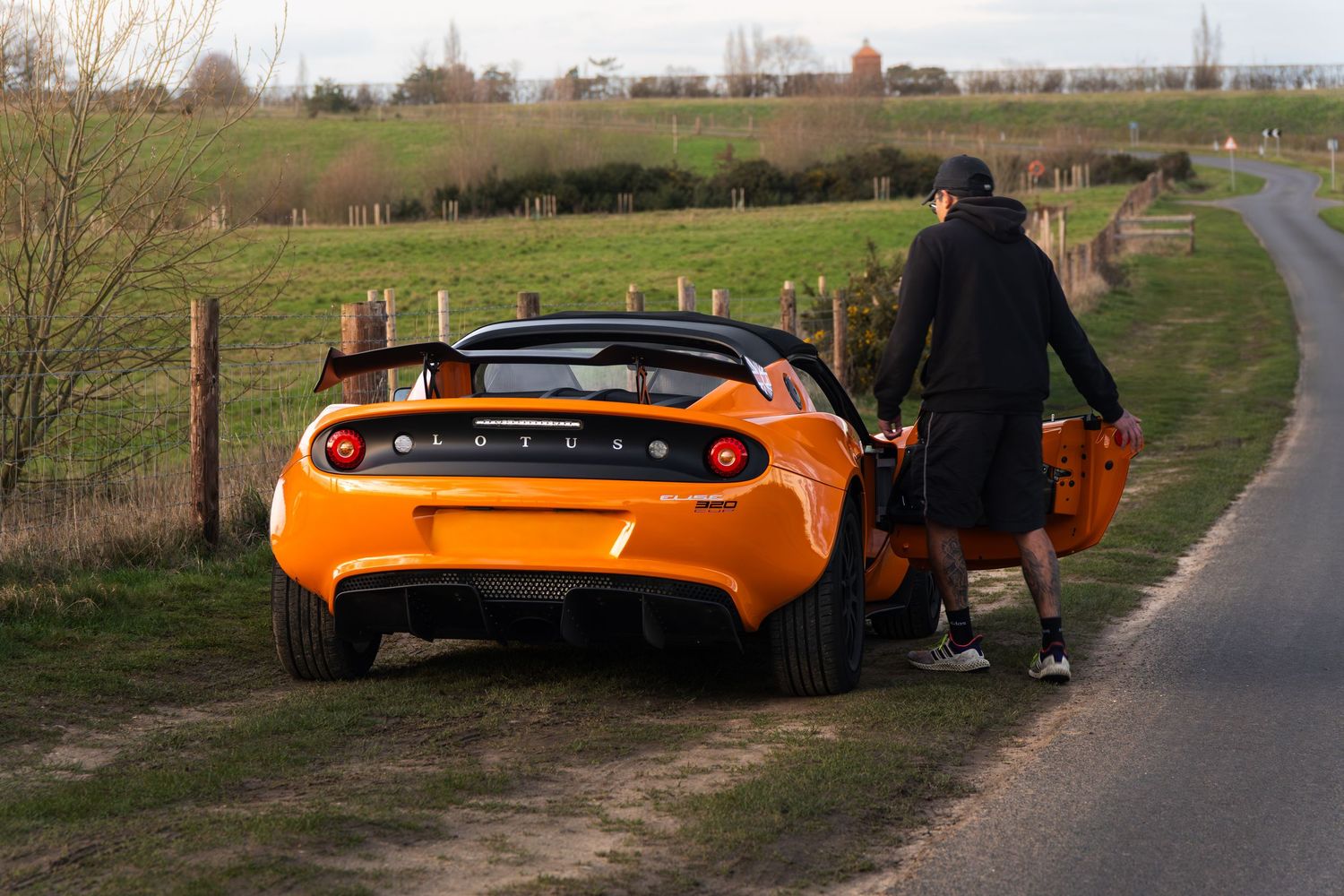 Person opening door of bright orange Lotus Elise parked on quiet rural country lane.