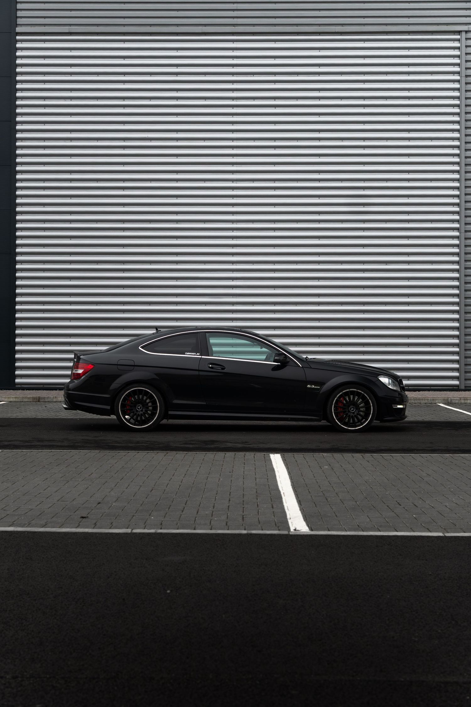 Black Mercedes-Benz C63 AMG coupe with red brake calipers parked against corrugated wall.