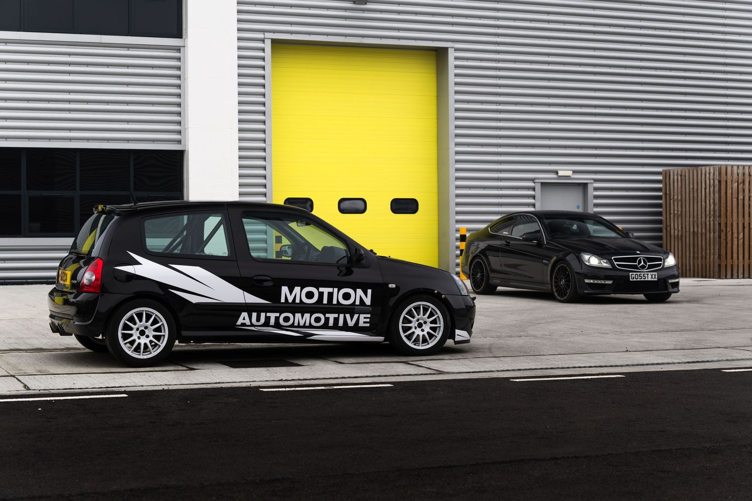 Black Motion Automotive Renault Clio and black Mercedes-Benz C-Class parked side by side outside yellow garage doors.