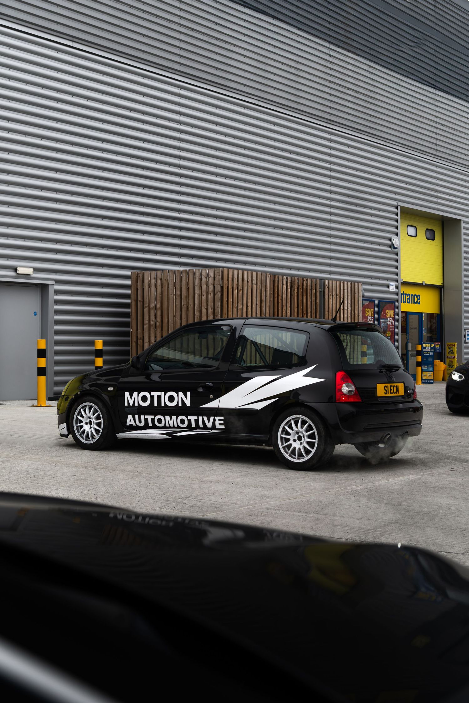 Side profile of a black Motion Automotive wrapped Renault Clio parked outside a grey industrial building.