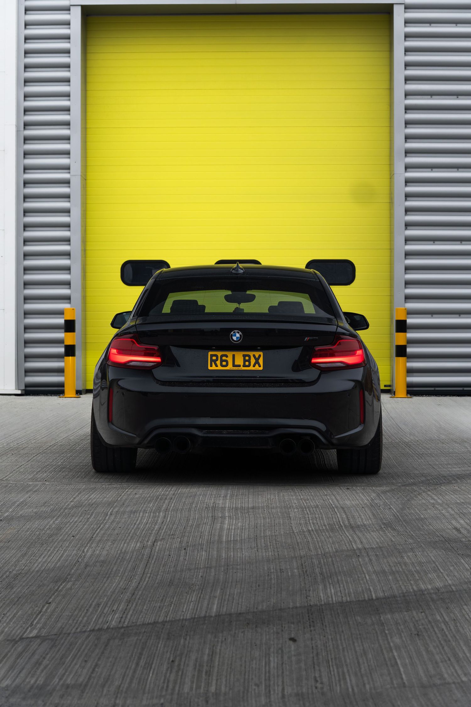Rear view of a black BMW M2 with plate R6LBX in front of a bright yellow roller door.
