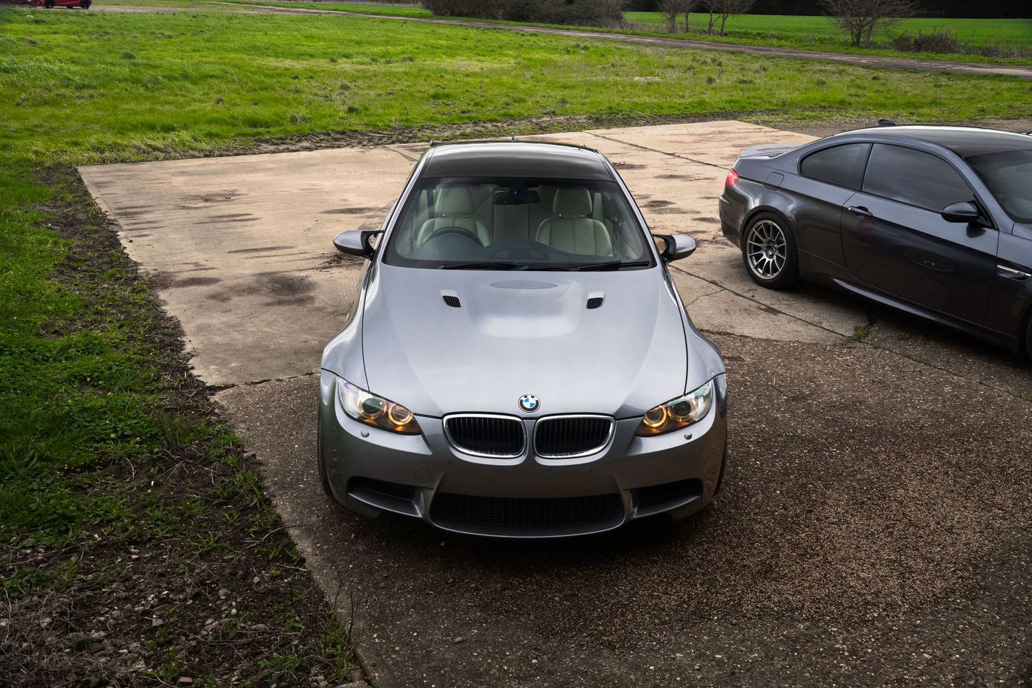 Front view of silver BMW M3 with angel eye headlights parked on concrete driveway near grass.