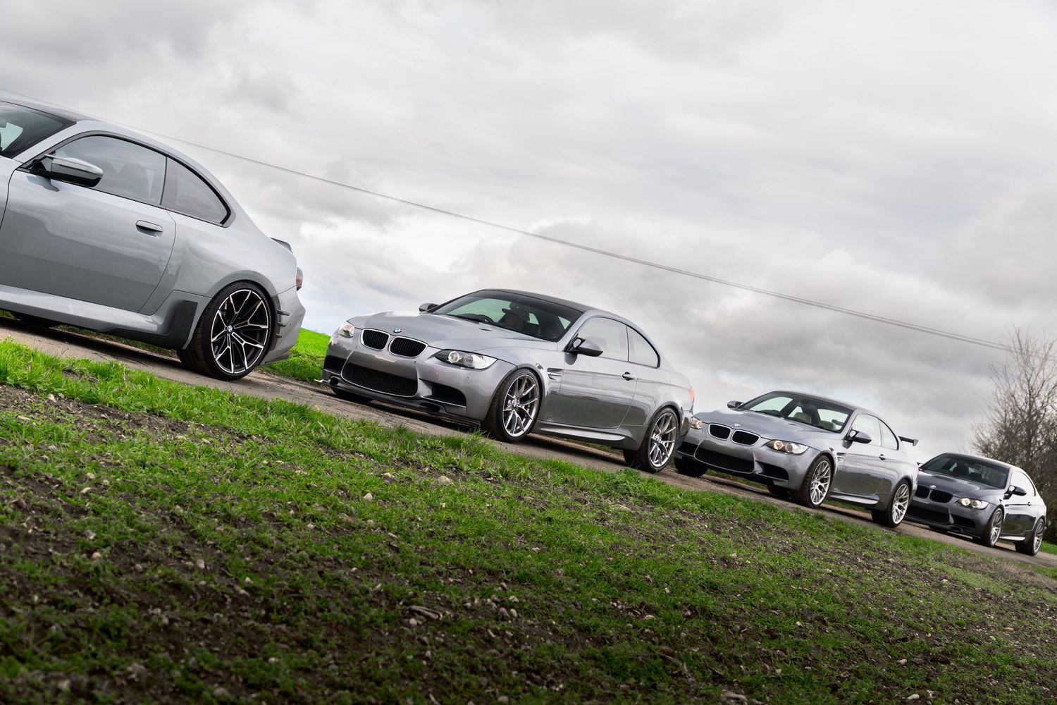 Four silver BMW M-series coupes lined up on a grassy rural road under dramatic overcast skies.