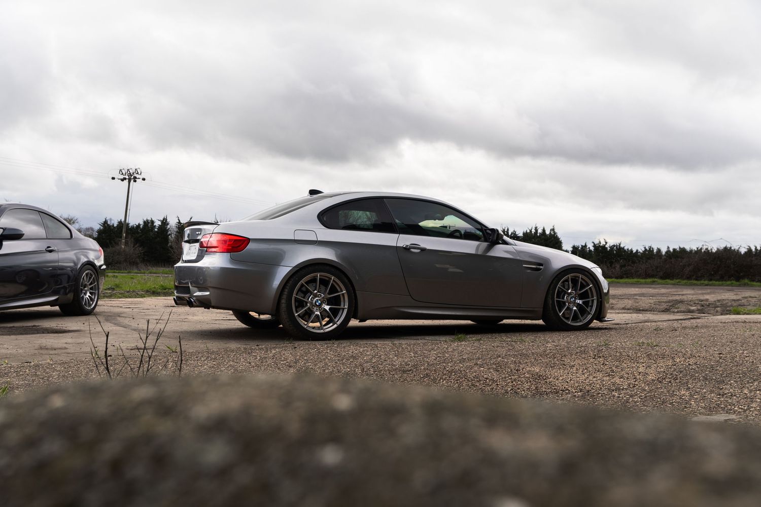 Side profile of a gray BMW M3 E92 coupe parked on gravel under an overcast sky.