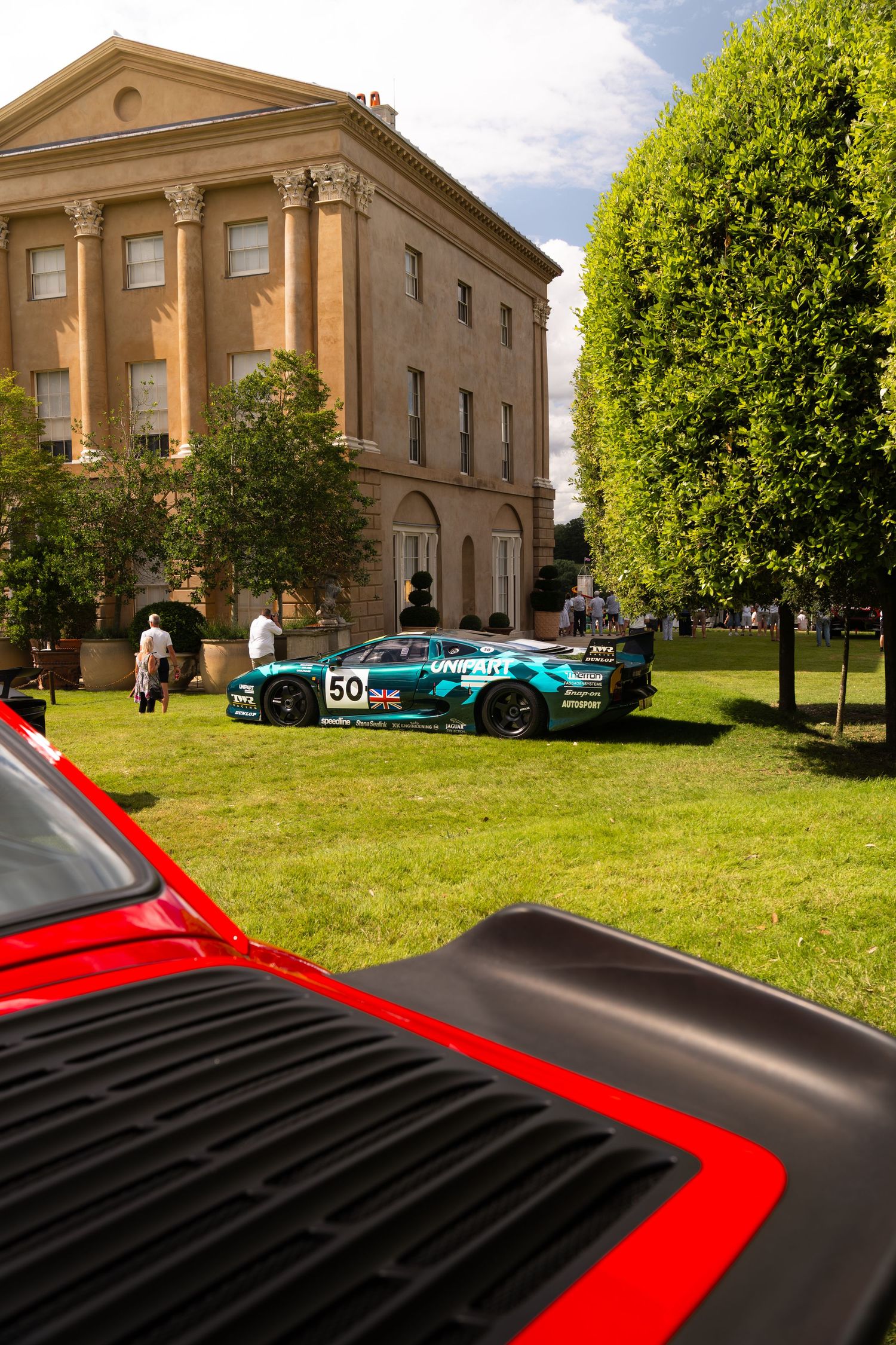 Teal race car #50 displayed on grass before a Georgian manor house at a classic car show, with a red sports car in foreground.