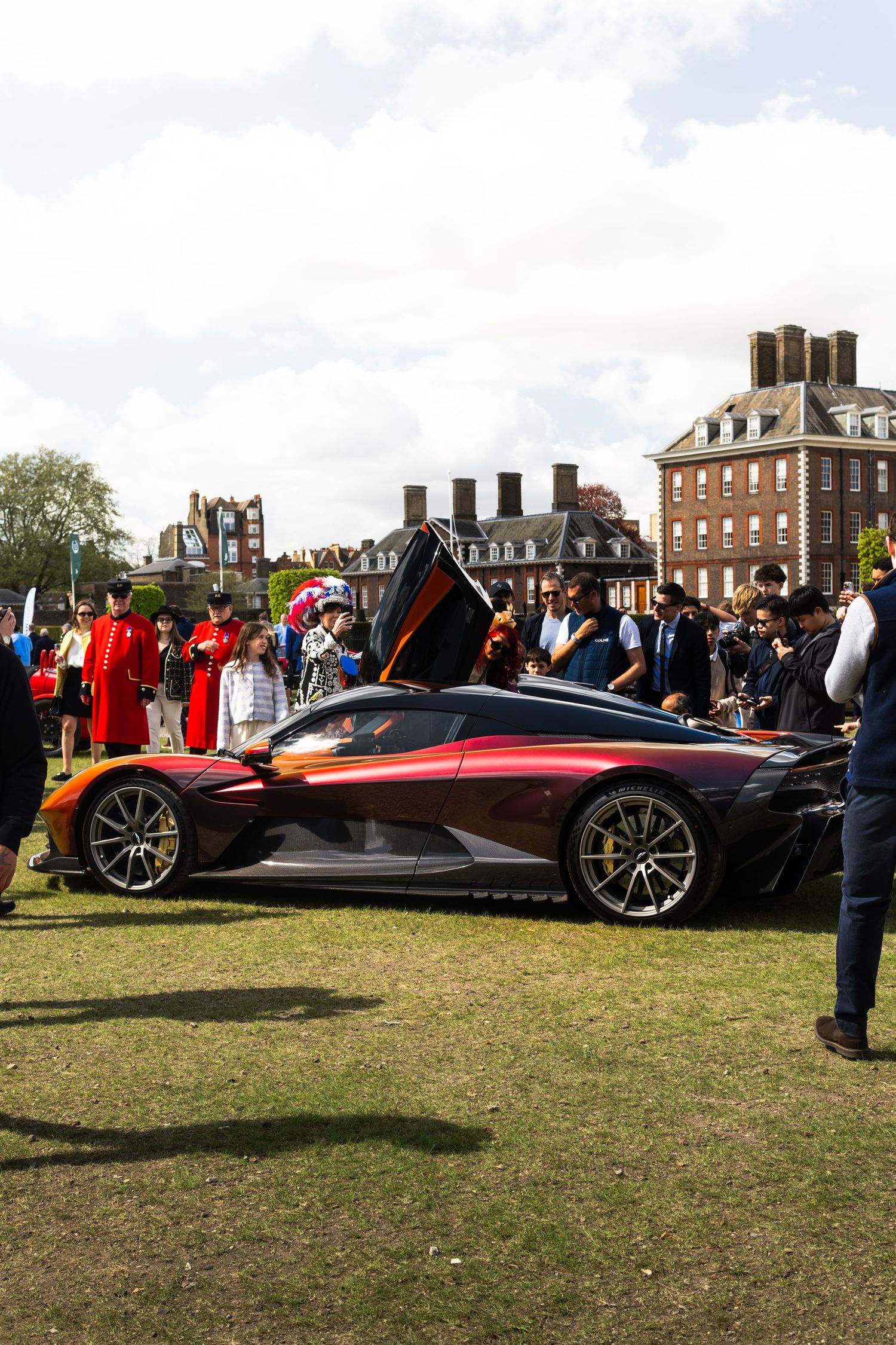 Dark hypercar with dihedral doors open displayed to crowds at an outdoor luxury automotive event.