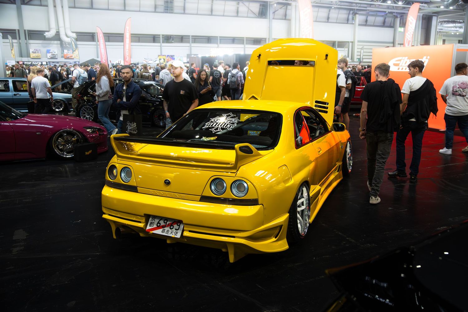 Yellow Nissan Skyline R33 GT-R with wide body kit on display at a car show with crowd of spectators.