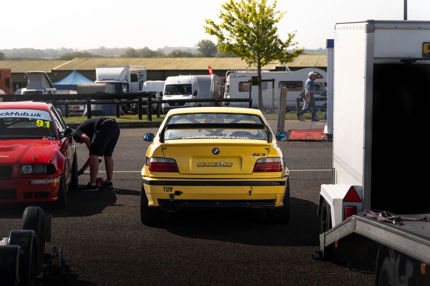 Yellow BMW E36 M3 race car viewed from rear in a motorsport paddock with trailers nearby.