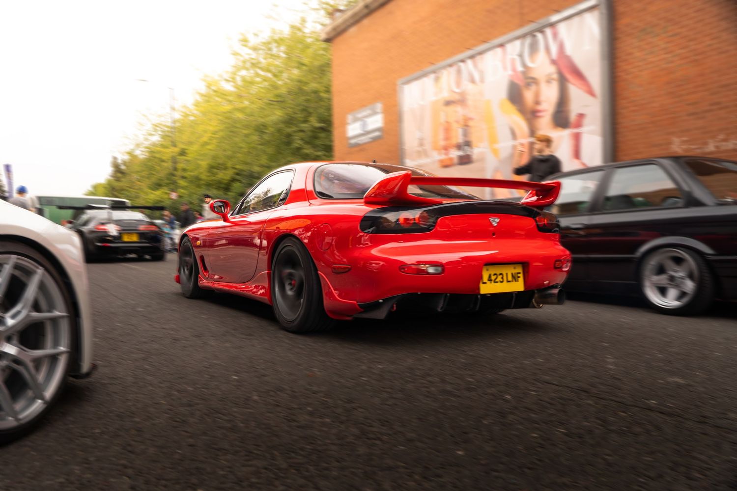 Red Mazda RX-7 with wide body kit and large rear wing at a car meet.