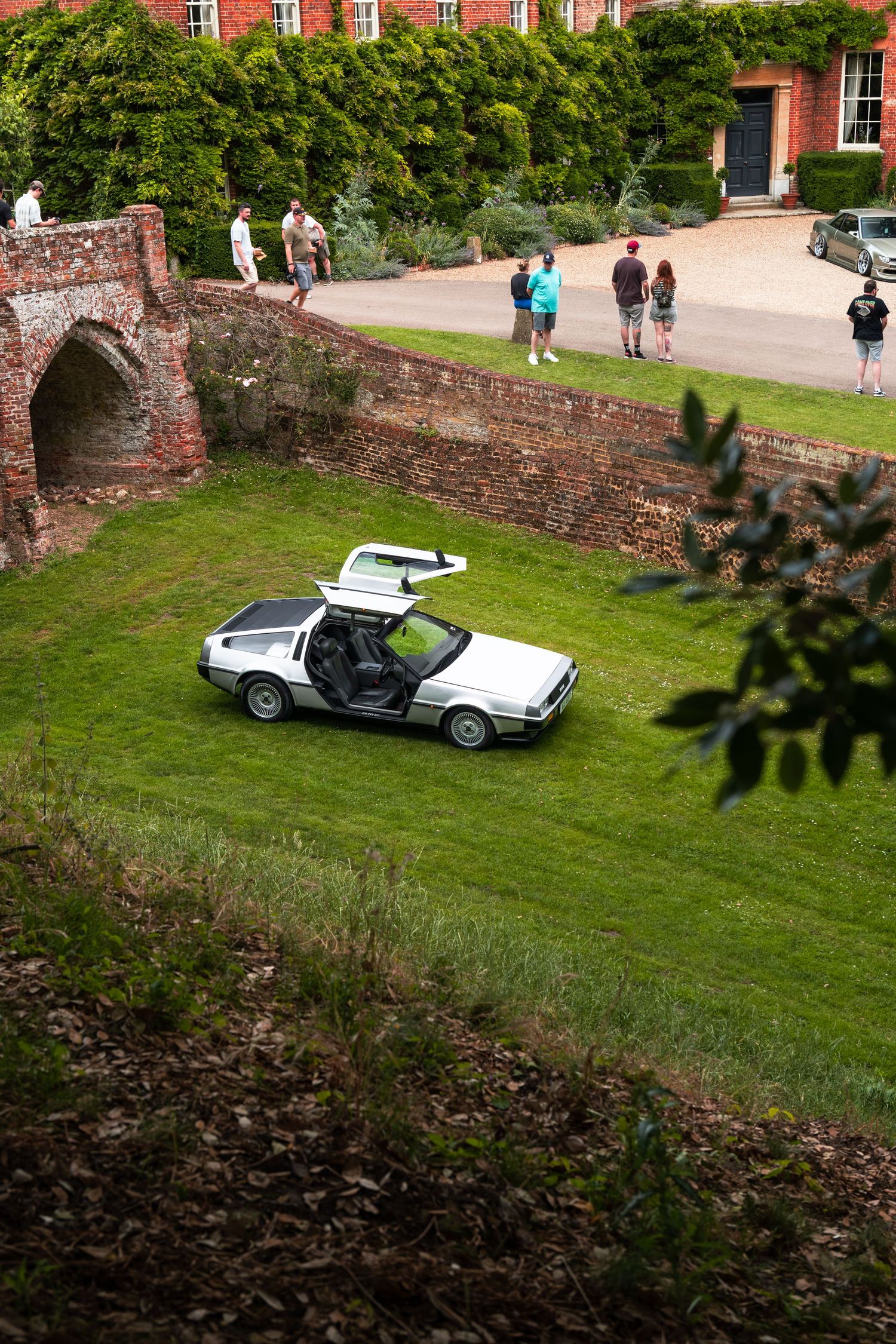 A silver DeLorean DMC-12 with gull-wing doors open sits on green grass beside a historic brick estate.