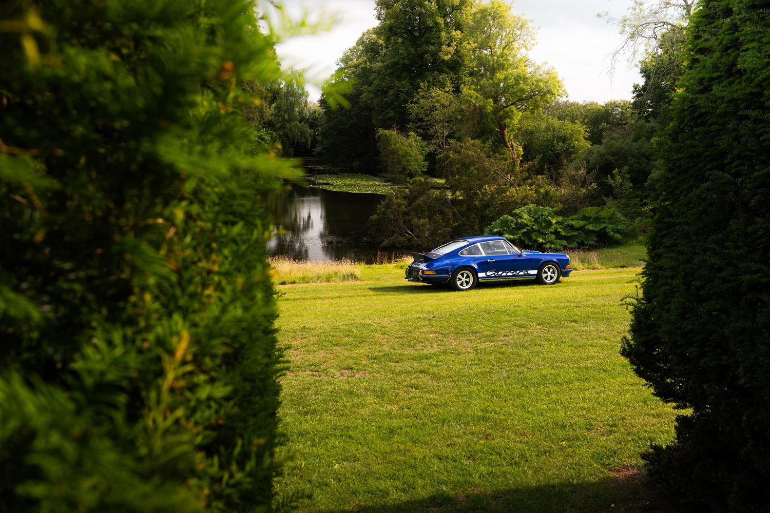Blue car parked on lush green lawn surrounded by trees with a pond and fountain in the background.