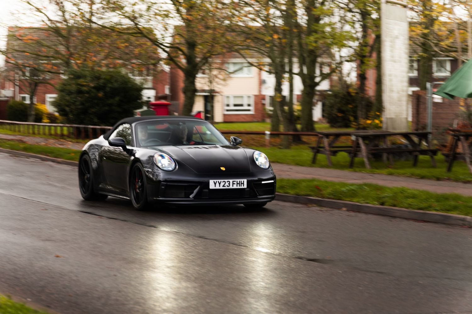 Black Porsche 911 Carrera driving on wet UK residential street on an overcast autumn day.