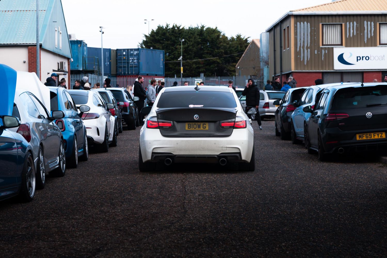 A white BMW 3 Series drives through rows of parked cars at an outdoor car meet.