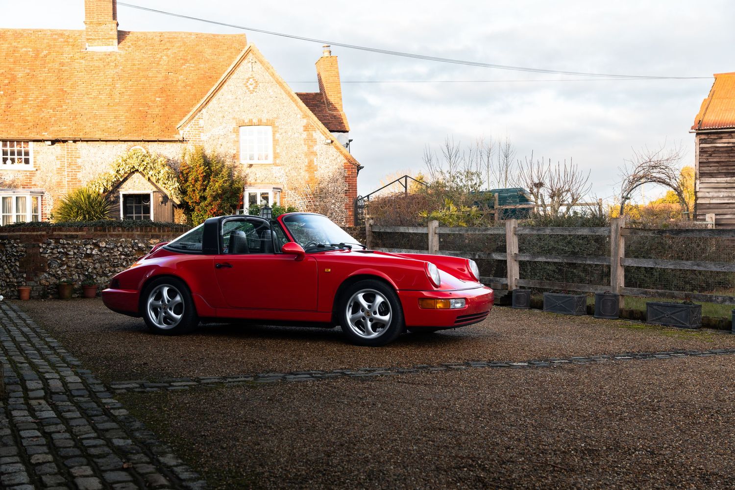 Vintage red Porsche 911 Carrera parked on gravel driveway beside a warm sunlit English rural stone building.