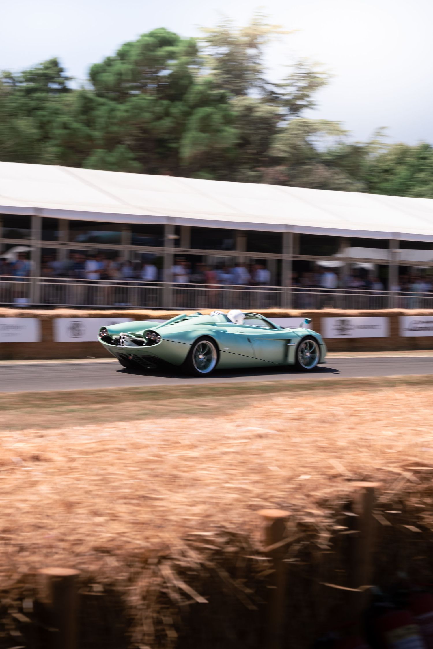 Vintage light blue sports car racing at Goodwood Festival of Speed, with spectators in grandstand behind hay bales.